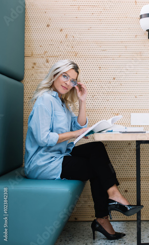 Portrait with side view of attractive smart female teenager in stylish optical eyewear holding literature book in hand and looking at camera while resting on publicity srea in college campus photo