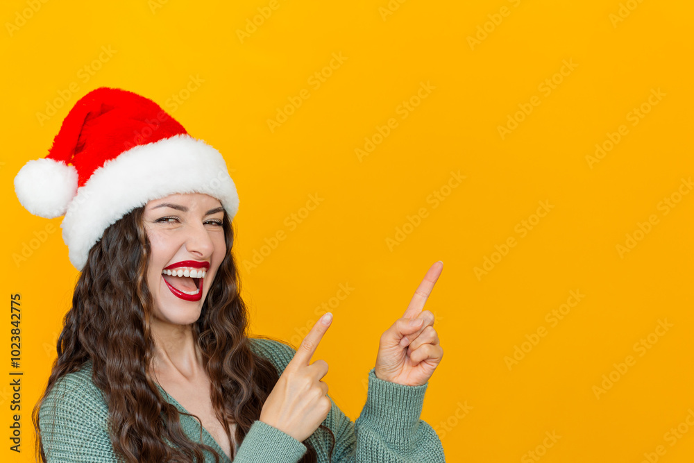 Young pretty brunette woman wearing Santa Claus hat and knitted green cardigan with red lipstick on her lips smiles with narrowed eyes and points her fingers at an empty background. Yellow background