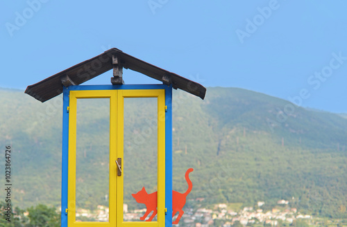 Fake red cat in bright yellow and blue window welcoming visitors in Brolo, the cat village at Lake Orta, with mountains in the back and blue sky on a summer day. Selective focus
