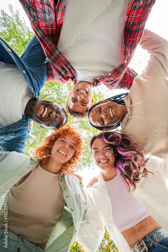 Vertical. Low angle view of a group of multiracial happy young teenagers hugging each others in a circle, looking down at camera with smiling faces representing friendship, diversity, and togetherness