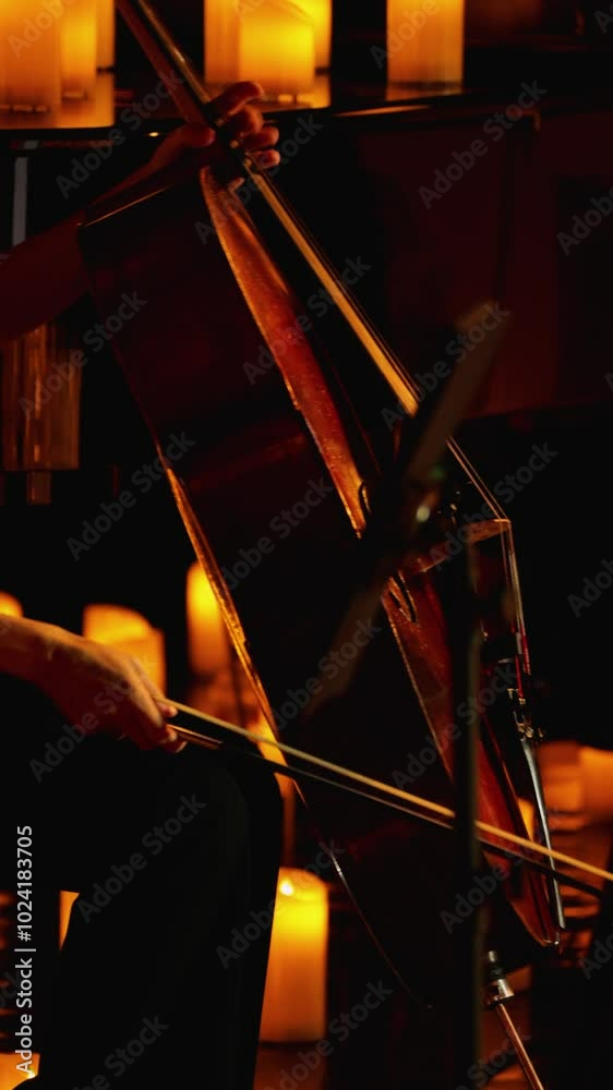 A graceful female hand with a bow plays the cello on a candlelit stage. The cello is illuminated by a warm light, highlighting the musician's hands as he plays