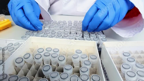 ab technician wearing gloves handles patient samples in test tubes in a genetic laboratory. The tubes are neatly arranged in a tray, indicating precise work with genetic material or blood diagnostics