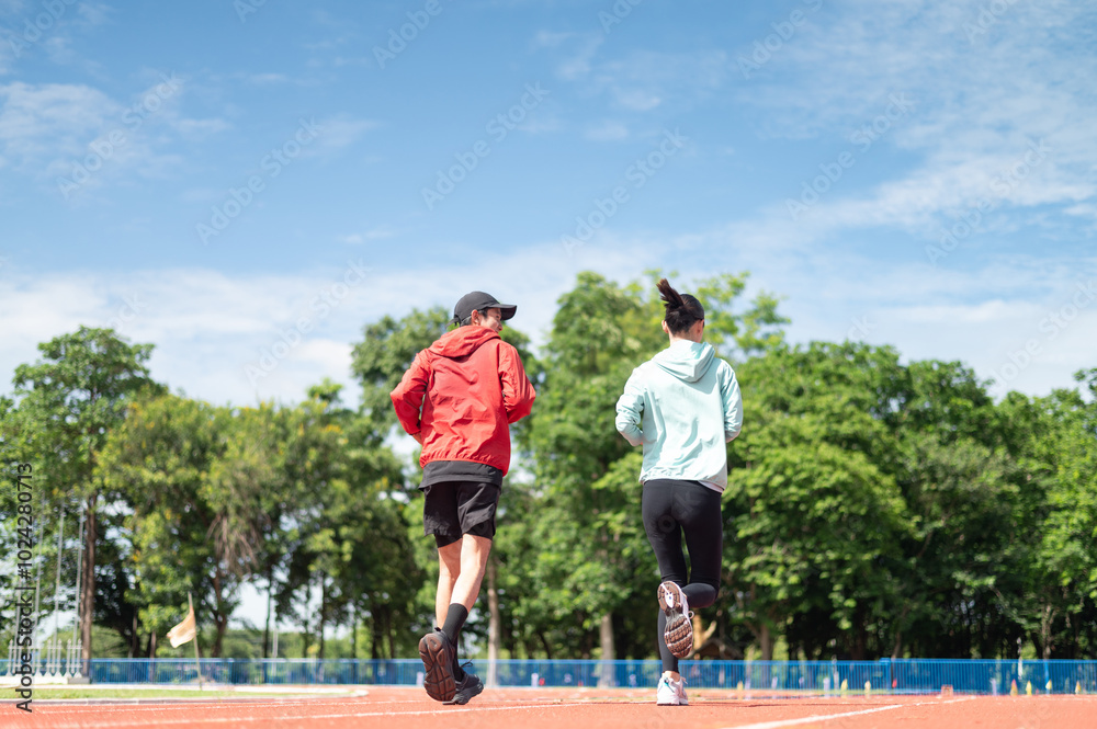Obraz premium A young Asian couple wearing blue and red sportswear, the two of them jogging together. Ready to talk while running outdoor exercise Exercise in the stadium maintaining health exercise active living