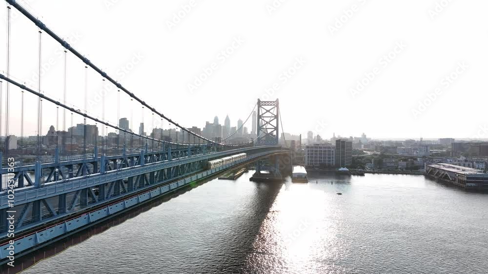 Aerial footage panning next to the Benjamin Franklin Bridge with the Patco trainline and the Philadelphia sunset skyline in the distance. Captured from the Camden Waterfront next to the Delaware River