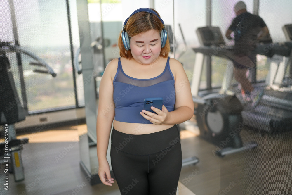 Fototapeta premium Obese asian woman Enjoy listening to music. Fat Female with headphones smart phone after workout at Gym