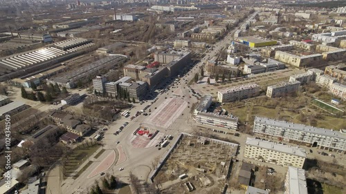 Aerial view of oval square in a city with low buildings. Cars drive along the ring road. There is a huge factory nearby.
