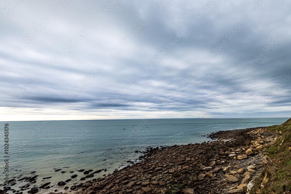 Serene cloudy coastal landscape featuring a rocky shoreline and calm ocean waves under a dramatic sky. Serene cloudy coastal landscape featuring a rocky shoreline and calm ocean waves under a dramatic sky.