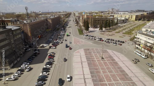 Aerial view of oval square in a city with low buildings and monument to Ordzhonikidze. Cars drive along the ring road. There is a huge factory nearby.