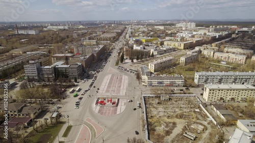Aerial view of oval square in a city with low buildings. Cars drive along the ring road. There is a huge factory nearby.