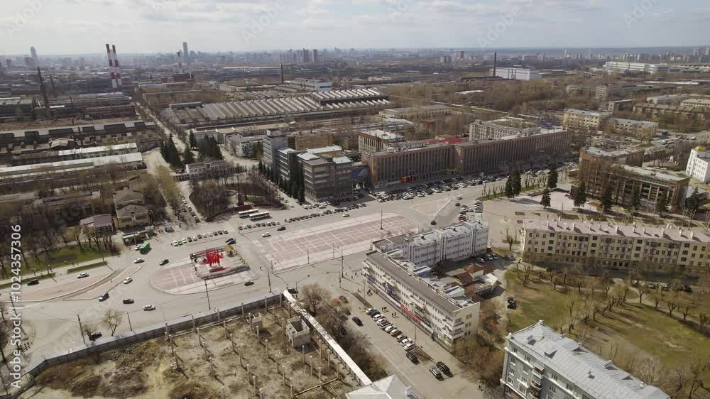 Aerial view of oval square in a city with low buildings. Cars drive along the ring road. There is a huge factory nearby. Aerial view of oval square in a city with low buildings. Cars drive along the ring road. There is a huge factory nearby.