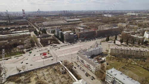Aerial view of oval square in a city with low buildings. Cars drive along the ring road. There is a huge factory nearby.