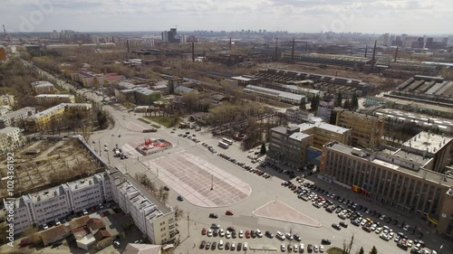 Aerial view of oval square in a city with low buildings. Cars drive along the ring road. There is a huge factory nearby.
