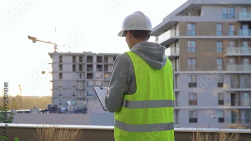 Man engineer with a white protective helmet and safety vest is writing on a clipboard while inspecting a construction site in early morning at sunrise. Architect concept