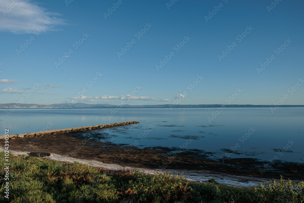 parte di spiaggia vicina, in primo piano, con vista della costiera di Trieste in lontananza, sotto un cielo sereno ed azzurro che si riflette sul mare Adriatico parte di spiaggia vicina, in primo piano, con vista della costiera di Trieste in lontananza, sotto un cielo sereno ed azzurro che si riflette sul mare Adriatico