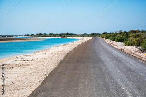 A long empty road runs along the shore of a turquoise lagoon. The road is surrounded by lush green vegetation and a bright blue sky