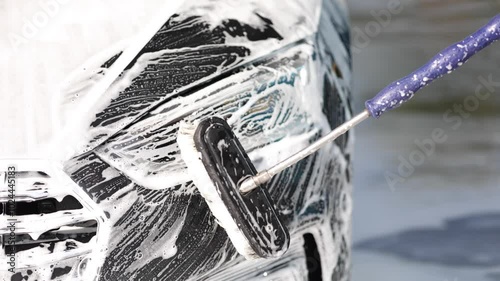 Man washes a car using a special brush at a car wash. Close up of man wash left headlight. The car is in foam