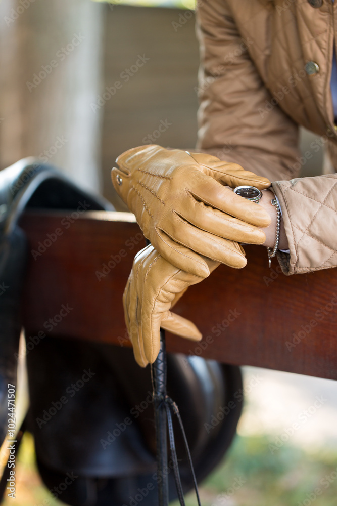 A woman in beige gloves looks at the clock while standing by a saddle in a stable