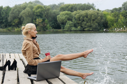 Woman office worker sitting on a pier by the river drinking coffee, talking on the phone and splashes water with his feet near the laptop