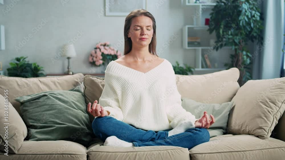 Woman wearing casual clothing is practicing yoga and meditating on a couch in her living room. The video is in slow motion Woman wearing casual clothing is practicing yoga and meditating on a couch in her living room. The video is in slow motion