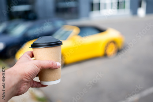 A hand holding a disposable coffee cup with a black lid, in focus. A blurred yellow sports car is visible in the background. Perfect for themes of travel, lifestyle, and on-the-go coffee culture.