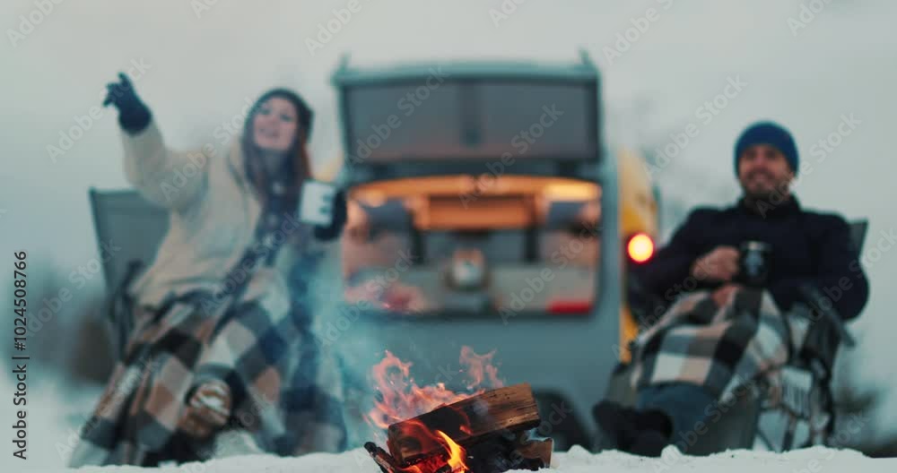 A couple is sitting by a campfire against the background of a camping on wheels on a winter, snowy day. The concept of outdoor recreation in a camping on wheels A couple is sitting by a campfire against the background of a camping on wheels on a winter, snowy day. The concept of outdoor recreation in a camping on wheels