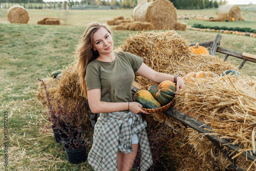 Young woman holding a basket of pumpkins near a haystack on a farm field. Outdoor autumn harvest scene. Fall season and agriculture concept for design and print photo