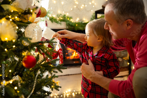 Father and his excited toddler celebrating Christmas at home. Family having fun decorating Christmas tree. Cute baby in red plaid pajamas. Beautiful house with fireplace, wrapped gifts on the floor.