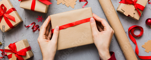 Woman is packing surprise gift for Christmas holidays, sitting at workplace with craft tools, top view. Woman wrapping Christmas gift at colored table