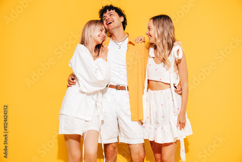 Group of young three stylish friends posing in studio. Fashion man and two cute female dressed in casual summer clothes. Smiling models having fun. Cheerful women and guy near yellow wall
