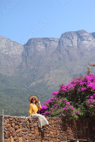 mulher sentada em muro ao lado de flores cor de rosa com serra do caraça ao fundo em catas altas, minas gerais  photo