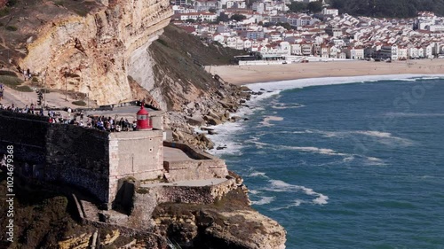 Aerial drone approaching the stunning cliffs and famous lighthouse at Nazare, Portugal during a sunny day at the beach with the village of Nazare in the background. Nazare is famous for giant waves