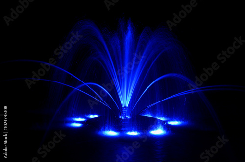 Floating fountain in neon blue, large detailed colorful horizontal closeup at night, lit up illuminated color changing bright city park lights, isolated colored water jets on dark black background photo