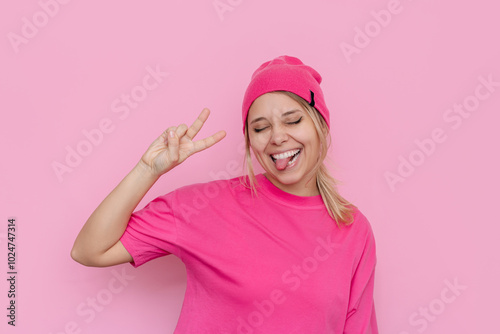 Stylish smiling young caucasian blonde woman with her eyes closed in a bright pink T-shirt and hat showing tongue and a peace gesture with her hand isolated on light pink color background