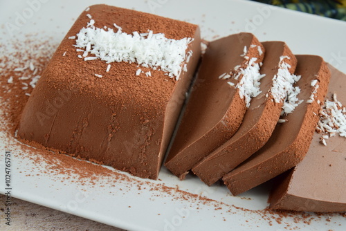 Dark chocolate milk pudding cake with cocoa powder and coconut flakes topping served on a plate. Selective focus