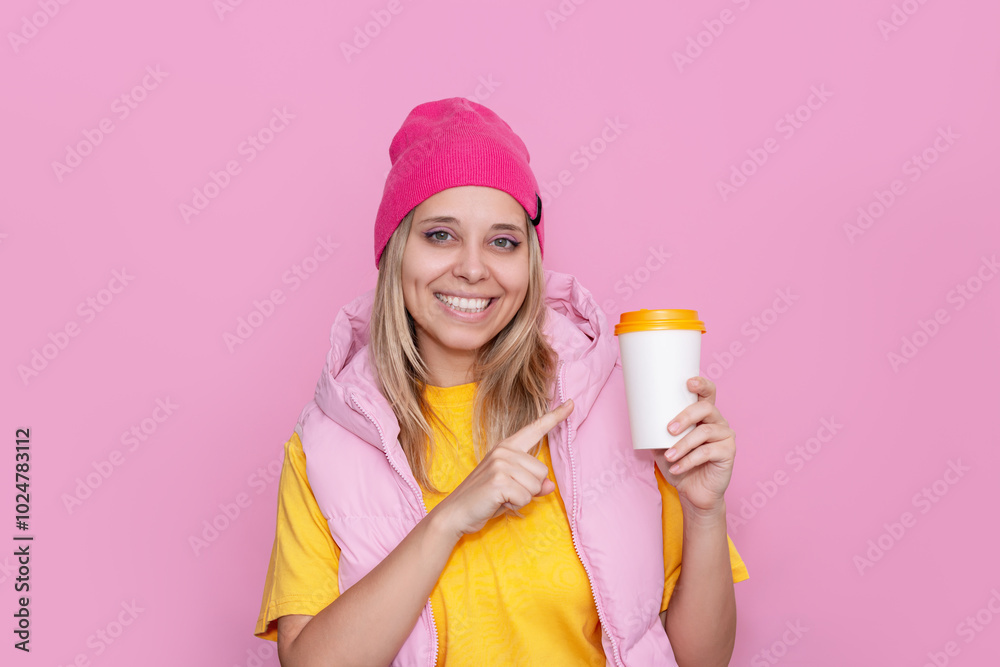Young smiling blonde woman in a yellow T-shirt and bright pink hat holding a white paper cup of coffee or tea pointing at empty copy space for text or design on a light pink color background Young smiling blonde woman in a yellow T-shirt and bright pink hat holding a white paper cup of coffee or tea pointing at empty copy space for text or design on a light pink color background