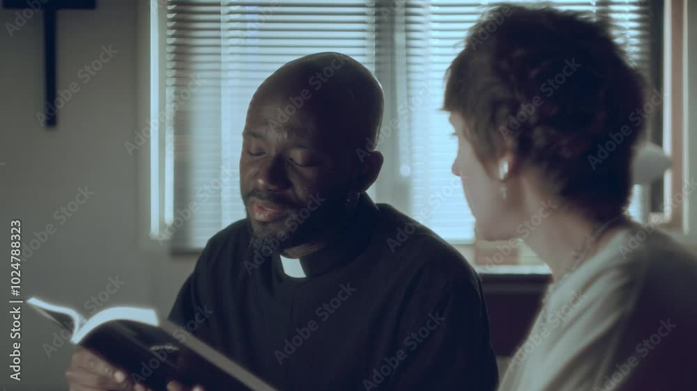 Black priest reading Holy Bible to young Caucasian woman in the living room at home, she sitting next to him on sofa and listening attentively. Rack focus