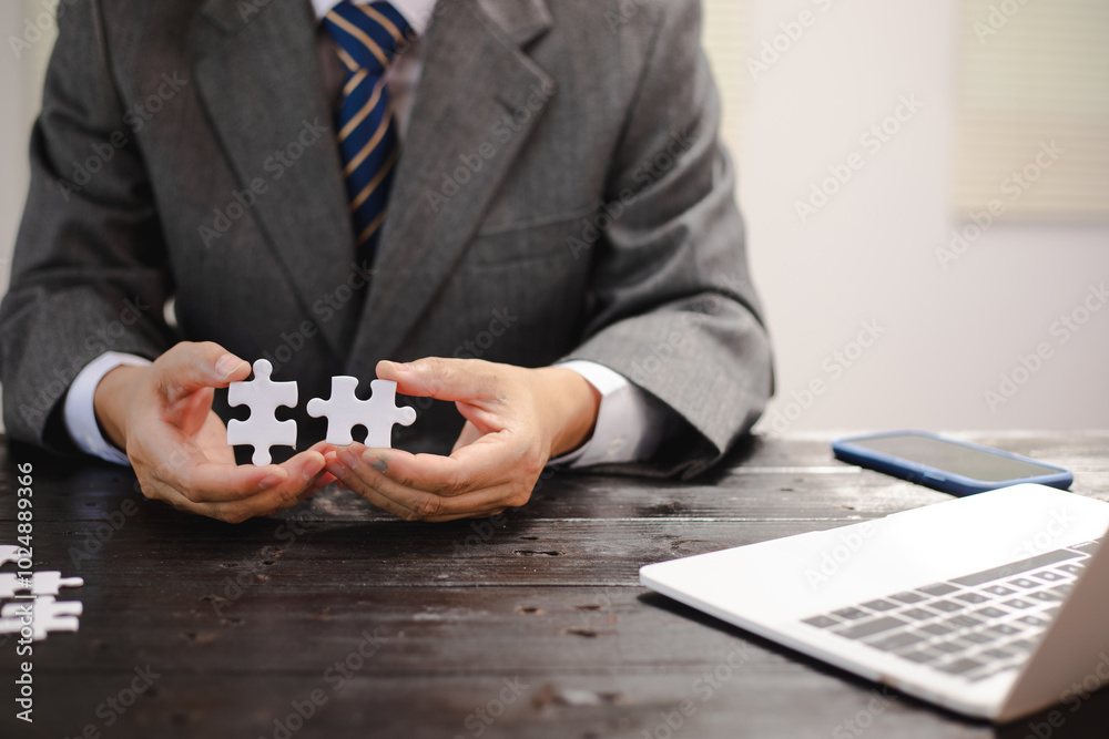 Businessman sitting at a table intently completing a jigsaw puzzle, demonstrating problem-solving, strategic thinking, and business decision-making, as well as attention to detail and patience. Businessman sitting at a table intently completing a jigsaw puzzle, demonstrating problem-solving, strategic thinking, and business decision-making, as well as attention to detail and patience.