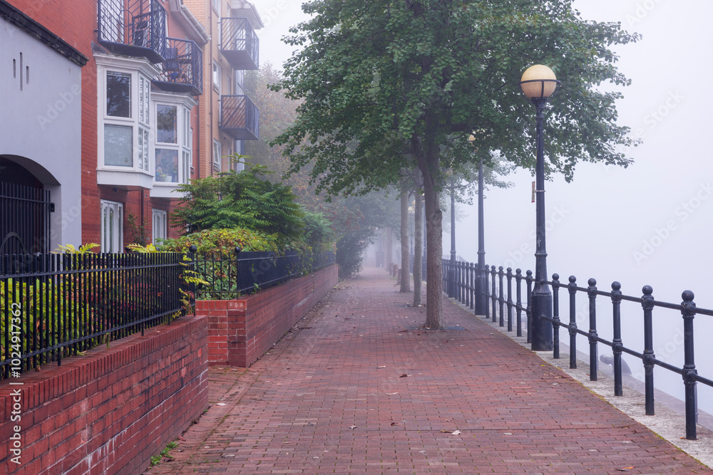Obraz premium Foggy Morning Along a Quiet Riverside Path Lined with Brick Apartments, Trees, and Classic Streetlamps, Creating a Serene Urban Scene Disappearing into the Mist