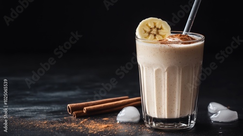 A creamy banana smoothie in a glass with ice cubes, cinnamon sticks, and a sprinkle of cinnamon on a black background.