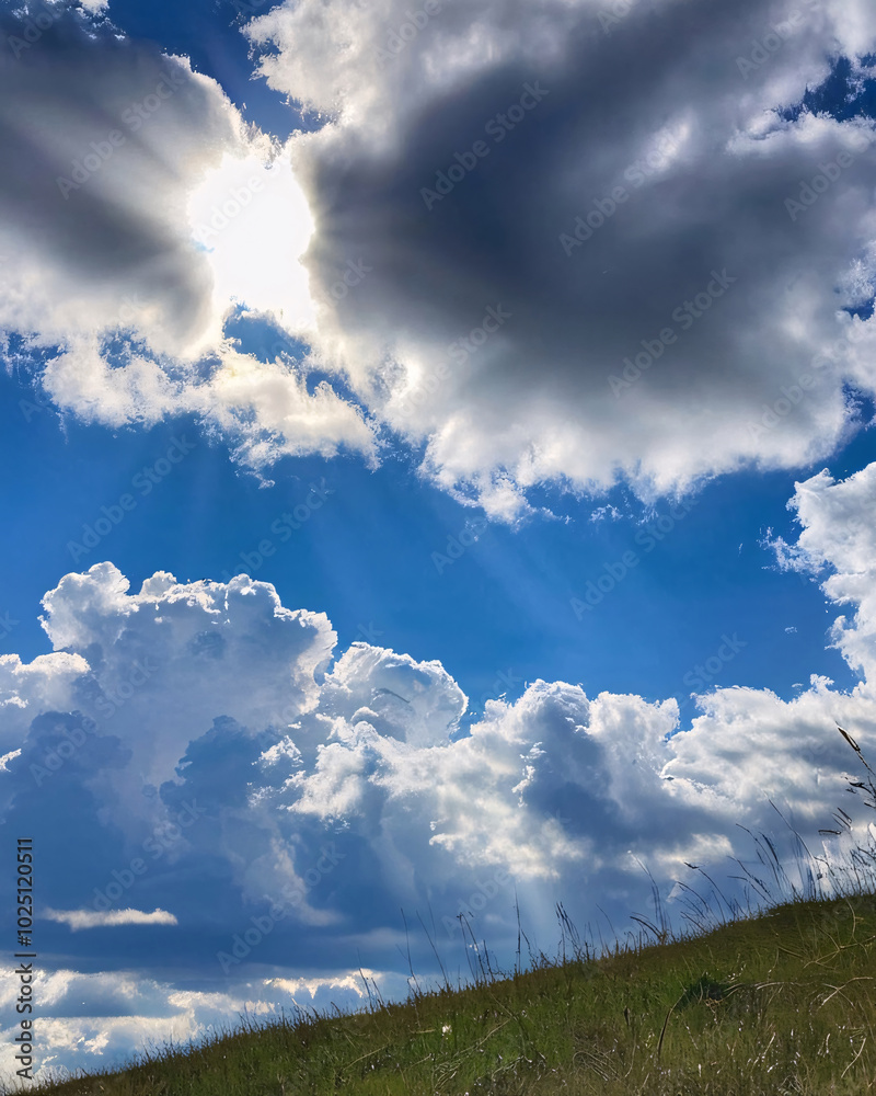 Bright white clouds contrasted against a clear blue sky, and sunlight shone through the clouds and down to the grassland below. Bright white clouds contrasted against a clear blue sky, and sunlight shone through the clouds and down to the grassland below.