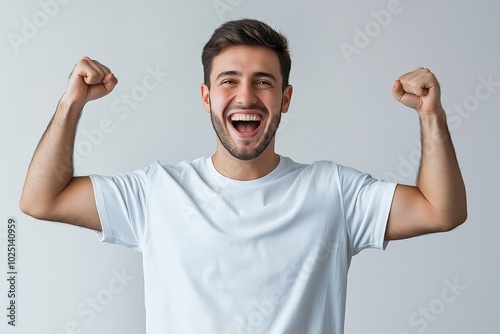 A Happy Young Man Celebrating Success in a Plain White T Shirt Against a Light Grey Background, Expressing Joy and Excitement With a Broad Smile and Raised Fists