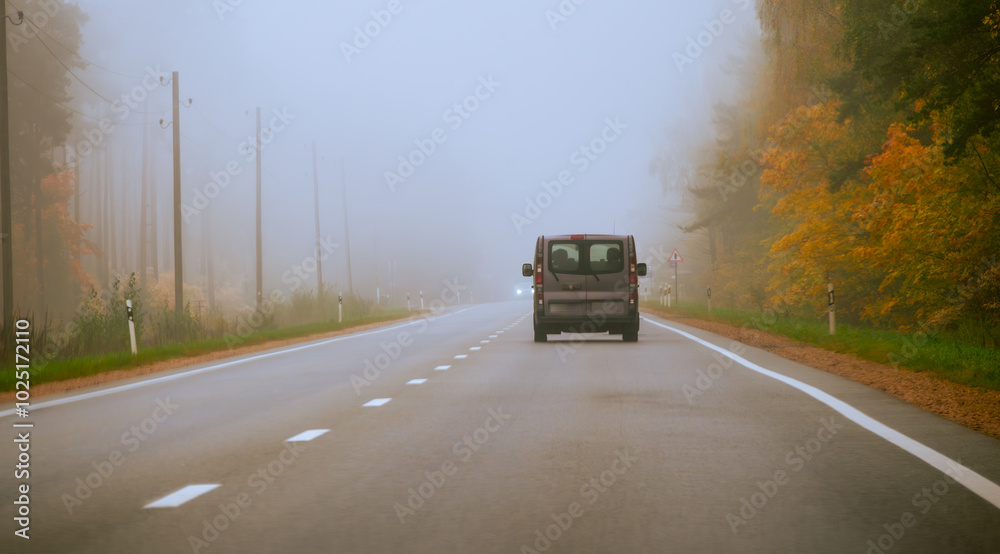 A Van Driving Along a Foggy Road Surrounded by Autumn Foliage in a Tranquil Rural Setting During the Early Morning Hours A Van Driving Along a Foggy Road Surrounded by Autumn Foliage in a Tranquil Rural Setting During the Early Morning Hours