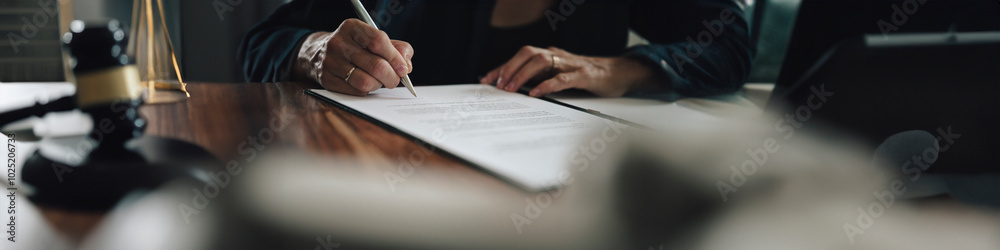 A person signs an official document at a wooden desk, with a gavel and legal tools nearby, symbolizing a formal agreement or legal decision in progress. A person signs an official document at a wooden desk, with a gavel and legal tools nearby, symbolizing a formal agreement or legal decision in progress.