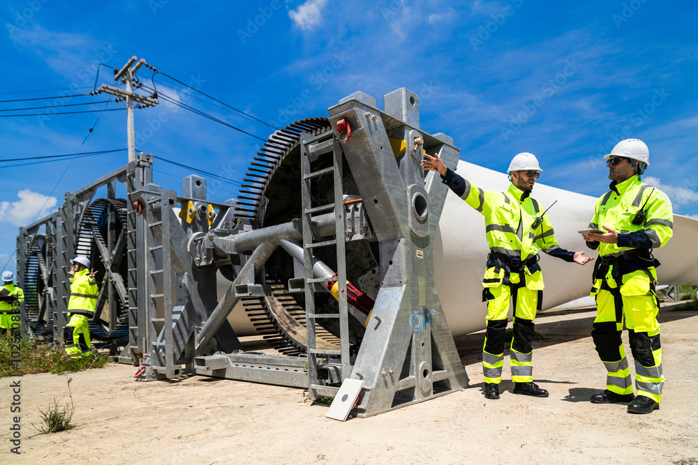 Fototapeta premium Two engineers in safety gear inspecting a wind turbine blade section on a construction site. They examine the metal framework with precision. Renewable energy project under a clear blue sky.