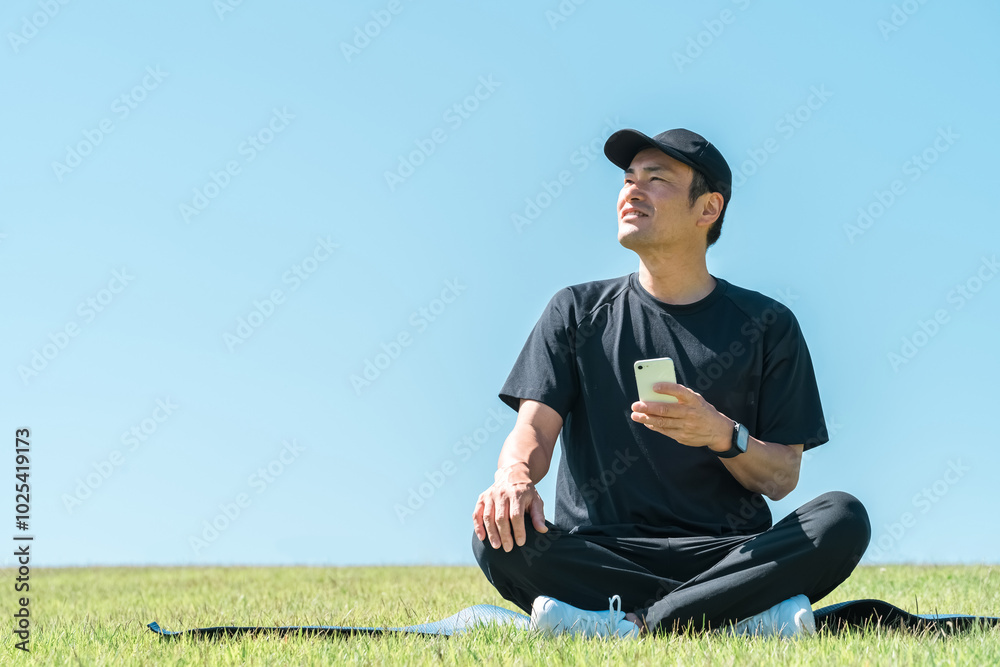 公園の芝生の上にヨガマットを引いて座りスマホを使うスポーツウェア姿のアジア人男性
公園の芝生の上にヨガマットを引いて座りスマホを使うスポーツウェア姿のアジア人男性