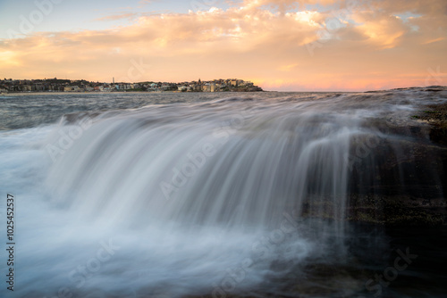 A gentle cascade flows over the rocky shore at Bondi Beach, blending with the soft hues of a pastel sunset, creating a serene coastal scene.