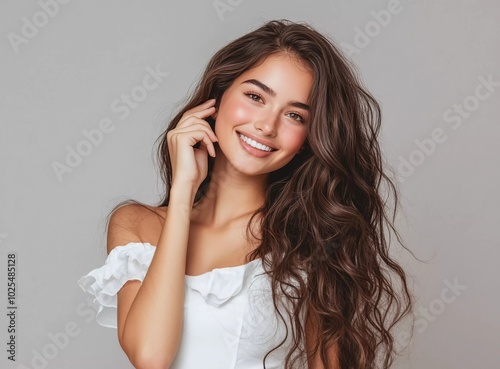 Beautiful woman with long brown hair in a white dress touching her curly hairstyle against a grey background