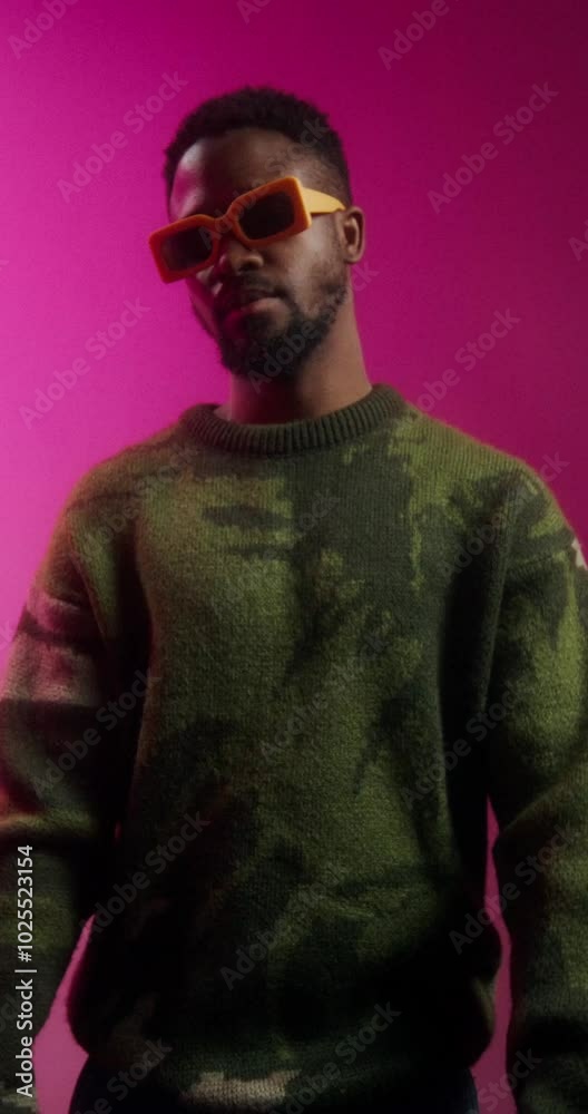 A bearded man in yellow sunglasses and a sweater poses standing on a pink background in a studio in the studio A bearded man in yellow sunglasses and a sweater poses standing on a pink background in a studio in the studio
