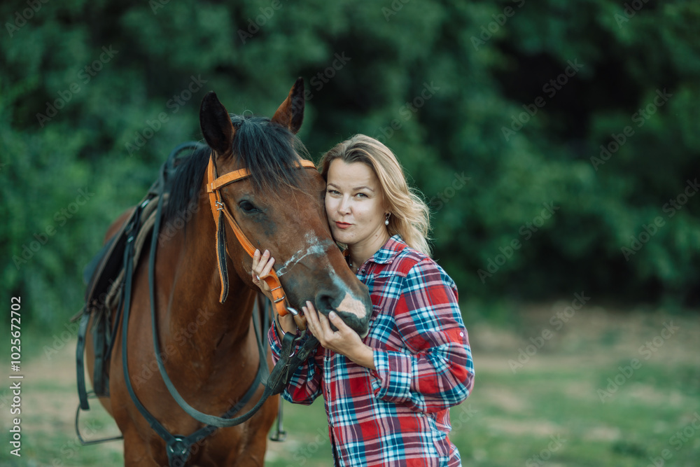 Obraz premium Happy blonde with horse in forest. Woman and a horse walking through the field during the day. Dressed in a plaid shirt and black leggings.