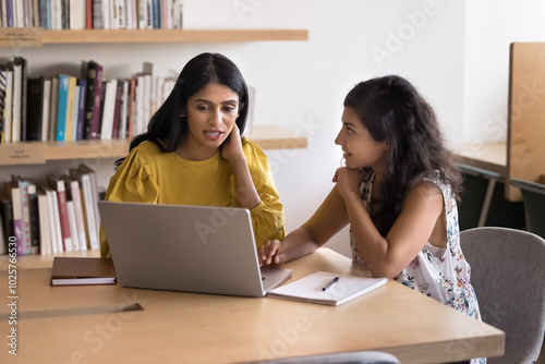 Two young Indian colleagues working on task together, talking at laptop on workplace table, watching, reading online content, discussing Intern application, software, website